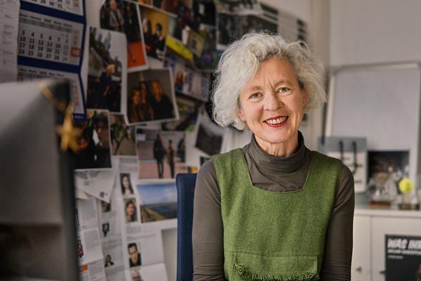 Inge sitzt vor ihrem Schreibtisch im Büro, hinter ihr zu sehen ist eine Pinnwand, die über und über mit Fotos bedeckt ist