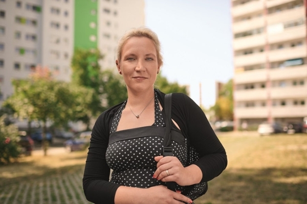 Mittelalte Frau mit blonden, zusammengebundenen Haaren, schwarz-weiß gepunktetem Kleid steht in einer Grünanlage einer Neubausiedlung