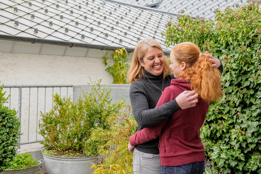 Mittelalte Frau mit rotblonden Haaren umarmt Teenagerin mit rotem Zopf im Garten vor einer Hauswand. Beide lachen bzw. lächeln.