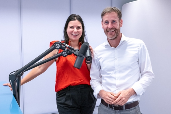 Eine Frau und ein Mann stehen an einem Tisch und unterhalten sich und sprechen in vor ihnen positionierte Mikrofone. Eine Frau und ein Mann stehen an einem Tisch und unterhalten sich und sprechen in vor ihnen positionierte Mikrofone.