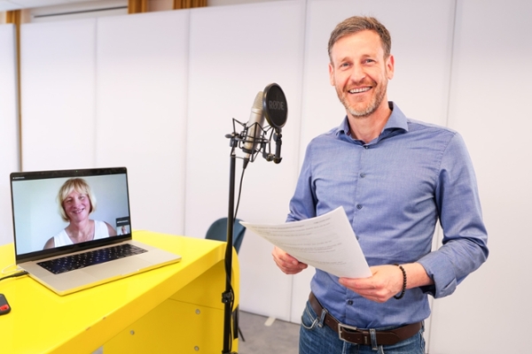 Ein Mann steht neben einem großen Mikrofon. Neben ihm ist ein aufgeklappter Laptop, auf dem eine Frau zu sehen ist. Ein Mann steht neben einem großen Mikrofon. Neben ihm ist ein aufgeklappter Laptop, auf dem eine Frau zu sehen ist.